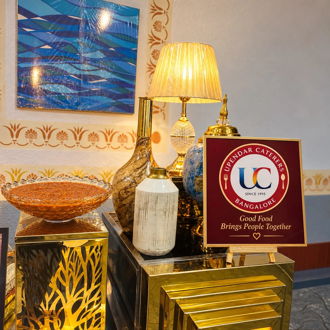 Brass buffet vessels beside a lamp and an Upendar Caterers sign